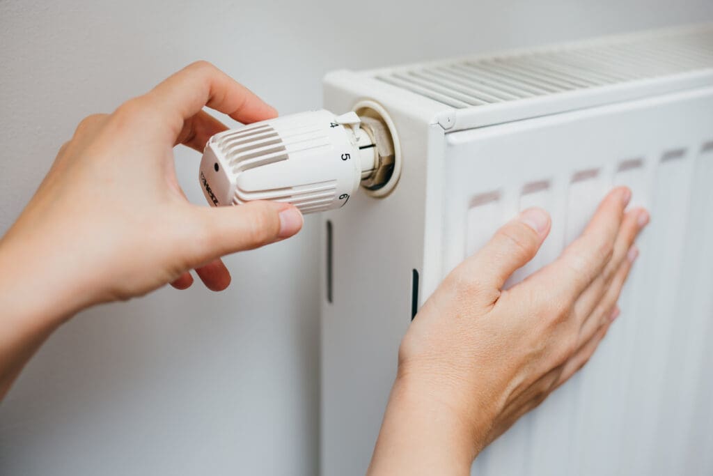 A person adjusts the temperature dial on a white radiator attached to a wall, using one hand to turn the knob and the other hand resting on the side of the radiator.