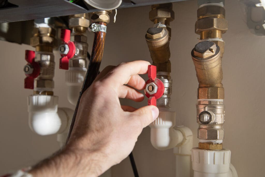 A person’s hand turning a red valve on a metal pipe among several plumbing pipes and fittings under a sink.