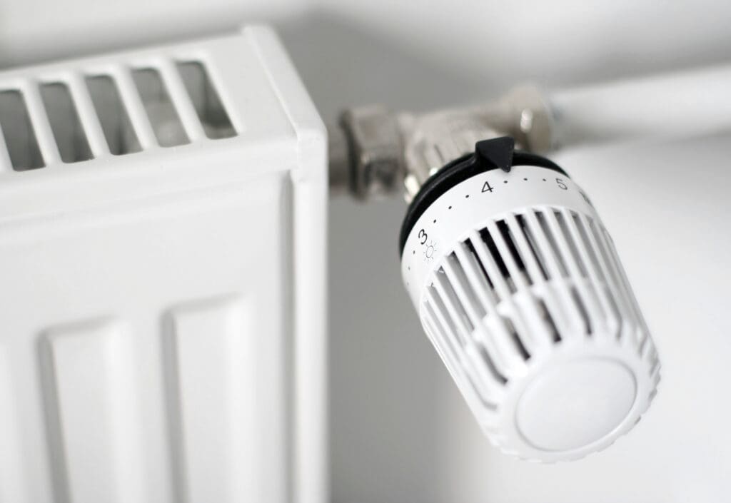 Close-up of a white radiator with a round, numbered thermostat dial attached, showing settings from 1 to 5. The dial is set between 3 and 4. The background and radiator are both white.