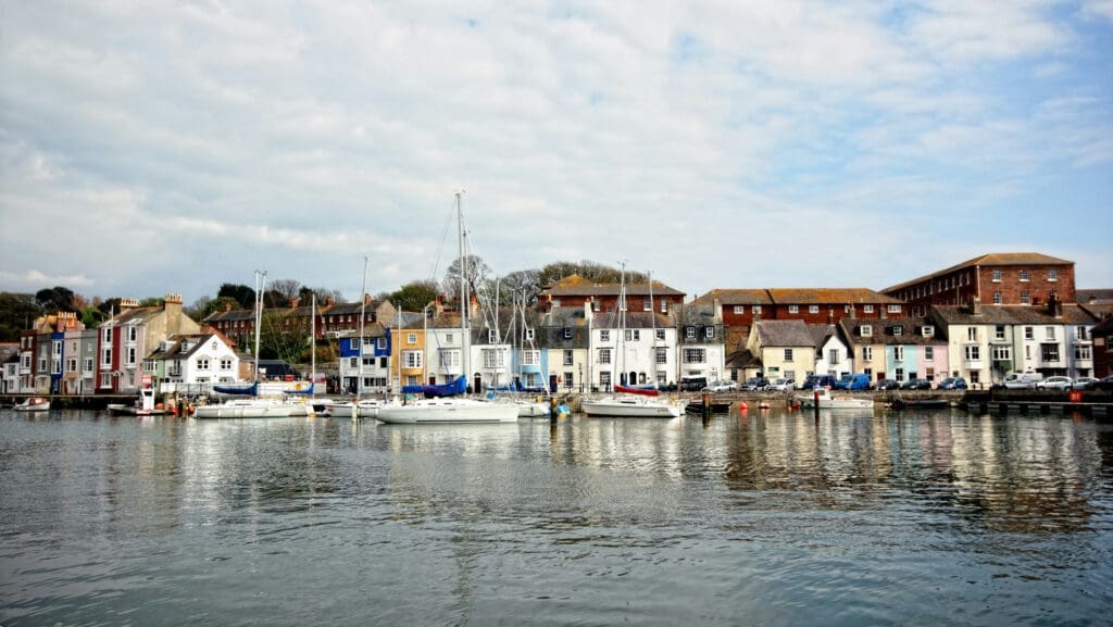 A row of colorful buildings and small boats line the waterfront on a calm, cloudy day, with reflections visible in the water and a few trees and brick structures in the background.