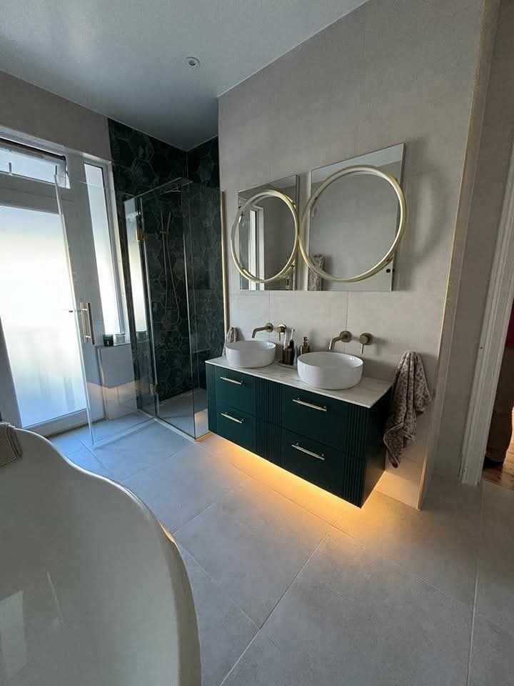 Modern bathroom with a glass shower, double sink vanity with vessel sinks, round mirrors with gold frames, and under-cabinet lighting. A bathtub edge is visible in the foreground.