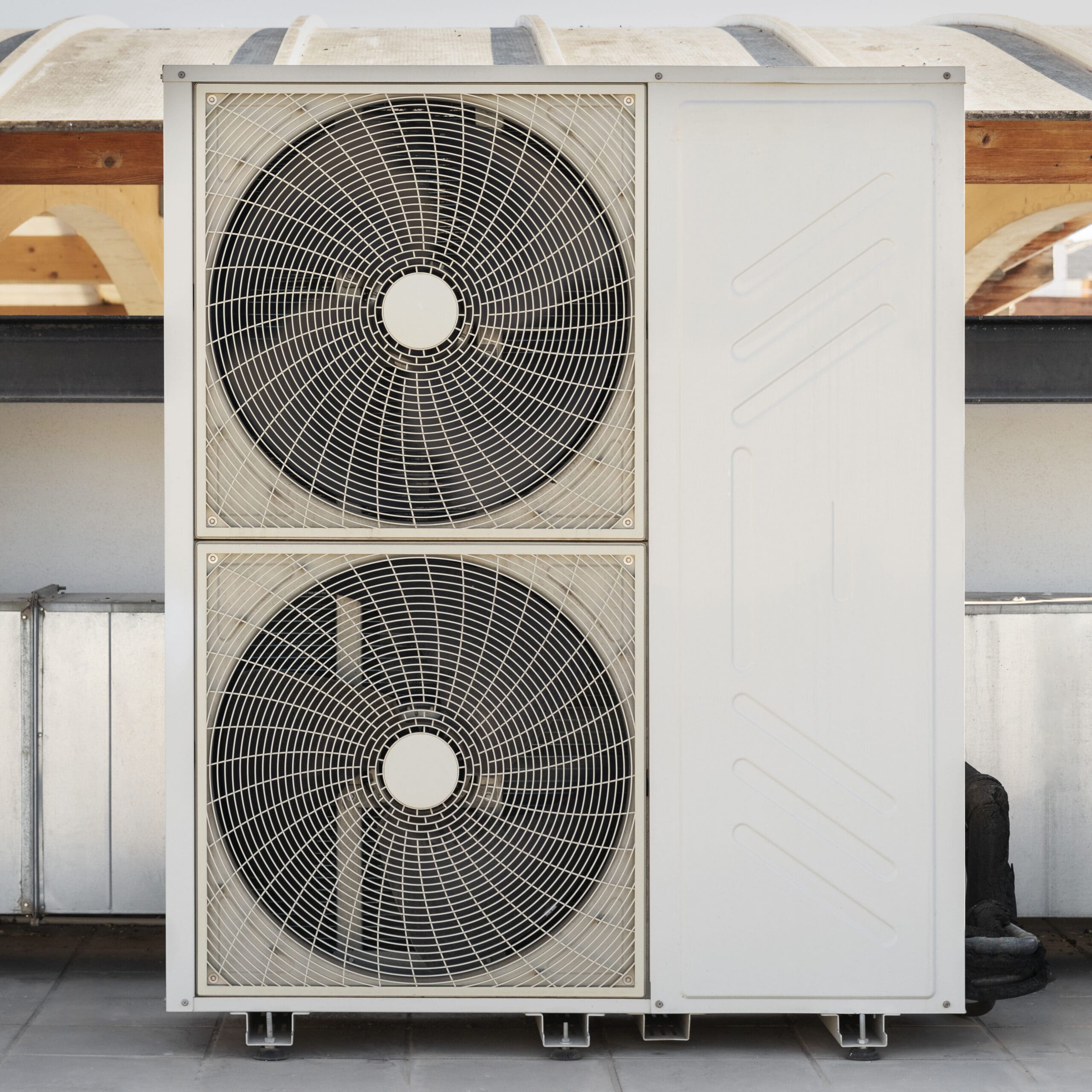 A large outdoor air conditioning unit with two circular fans stacked vertically, installed on a building rooftop with part of a wooden structure visible in the background.
