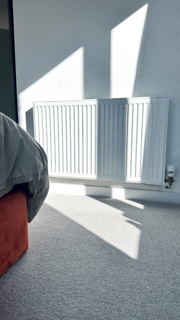 A white radiator mounted on a light grey wall, casting sharp sunlight and shadow patterns on the carpeted floor. Part of an orange bed and grey blanket are visible in the lower left corner.