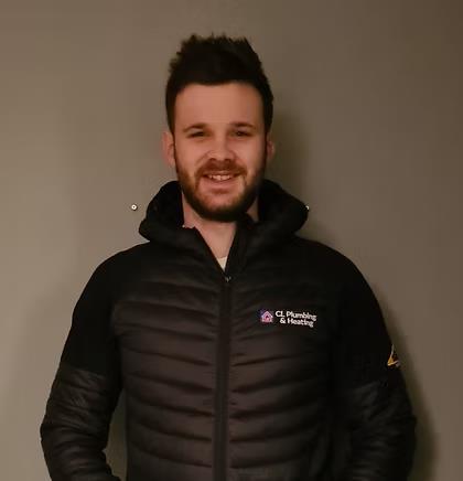 A man with short dark hair and a beard is standing and smiling, wearing a black jacket with the logo "CL Plumbing & Heating" on the chest, against a plain gray background.