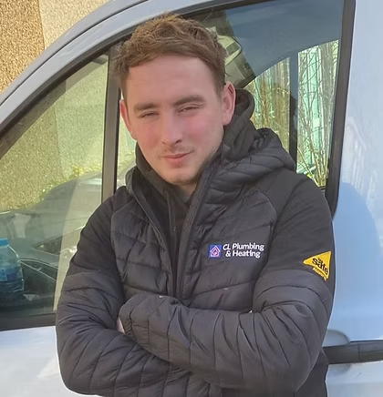 A man with short brown hair, wearing a black puffer jacket with "CL Plumbing & Heating" and "Safe" logos, stands in front of a white van with his arms crossed, looking at the camera.