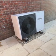 A white Vaillant heat pump unit is installed on a gravel bed next to a brick wall and surrounded by light-colored paving slabs.