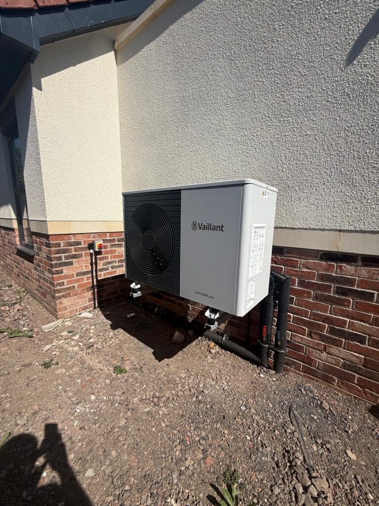A Vaillant heat pump unit is installed against the exterior wall of a house, which has a brick and stucco finish. Pipes are connected to the unit, and the ground around it is bare soil with some scattered stones.